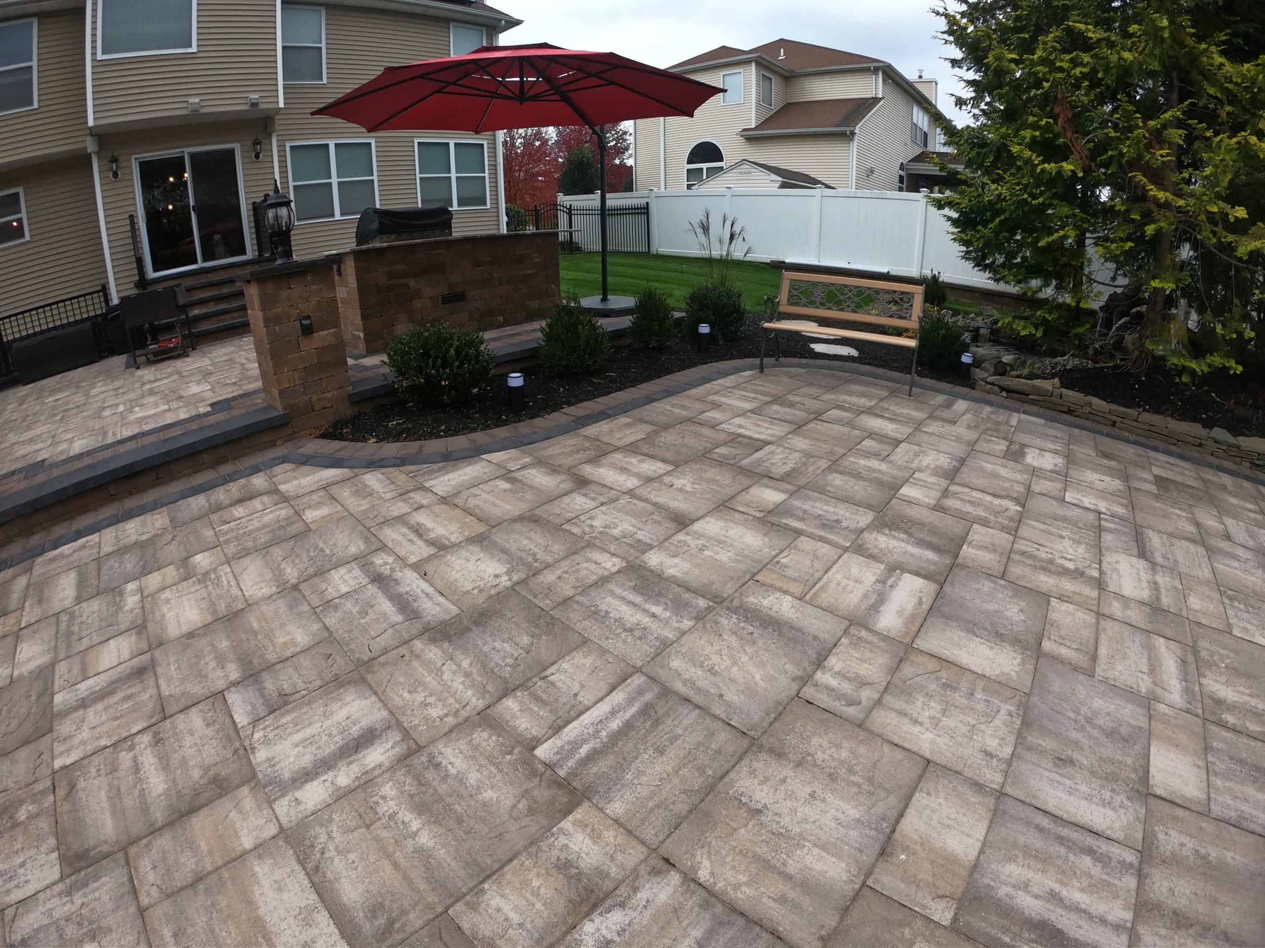 A spacious backyard patio with beige masonry pavers in Suffolk County, NY, features a bench and small garden area. An umbrella with a red canopy shades part of the seating area. The patio is adjacent to a house with large windows and a grass lawn bordered by a white fence.