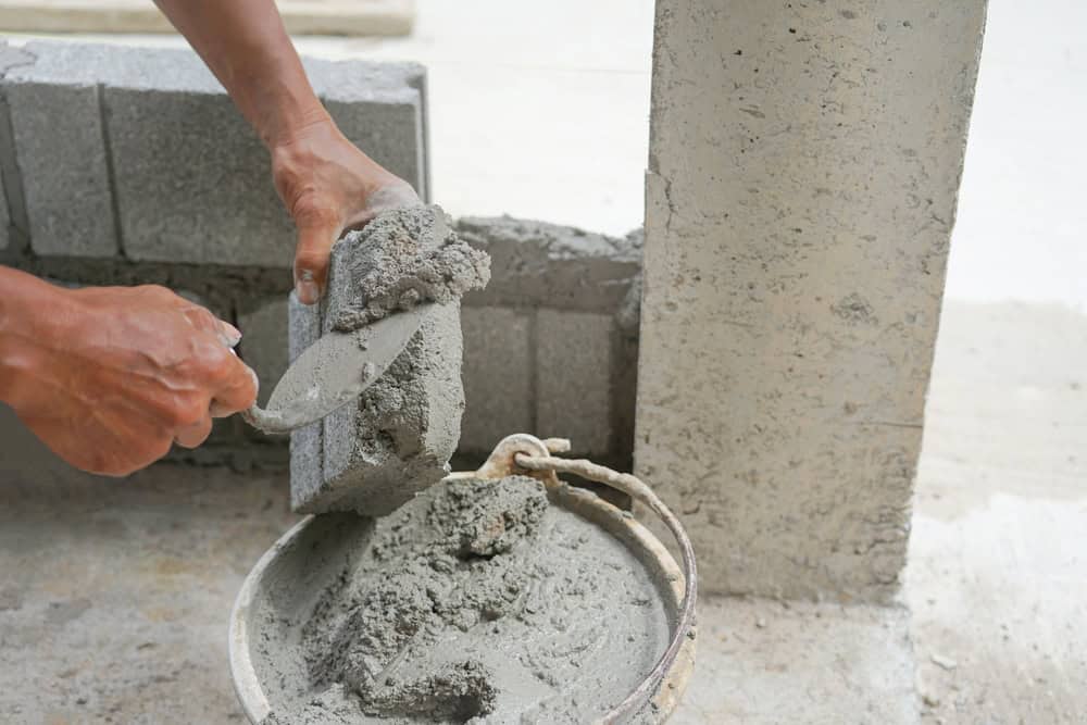 A person applies mortar to a concrete block using a trowel. The block is being prepared to add to a partially built wall. A bucket of mixed mortar is beside them on the ground. The scene is set at a construction site.