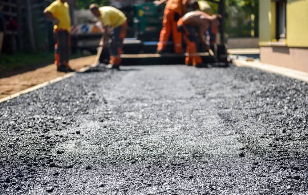 Workers in yellow and orange uniforms are paving a road with asphalt. The focus is on the freshly laid, rough-textured asphalt in the foreground, while the workers and paving machinery are slightly blurred in the background.