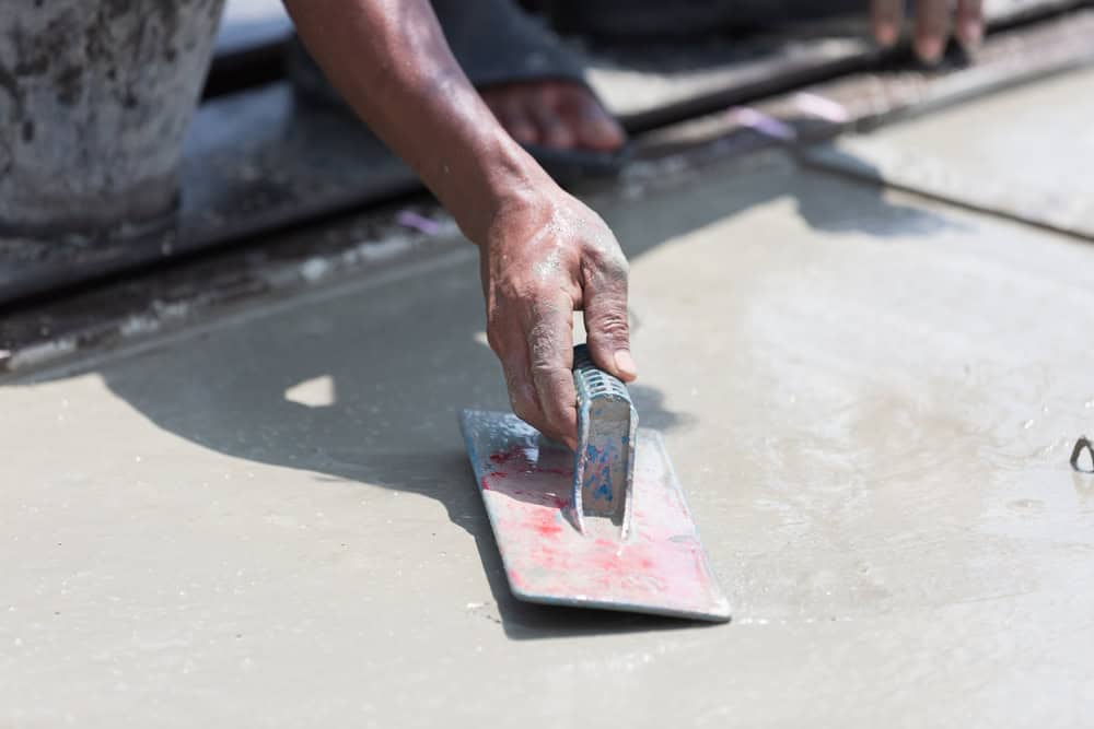 A person smoothing wet cement with a trowel. Their hand is steady as they work on the surface. The setting appears to be outdoors on a construction site.