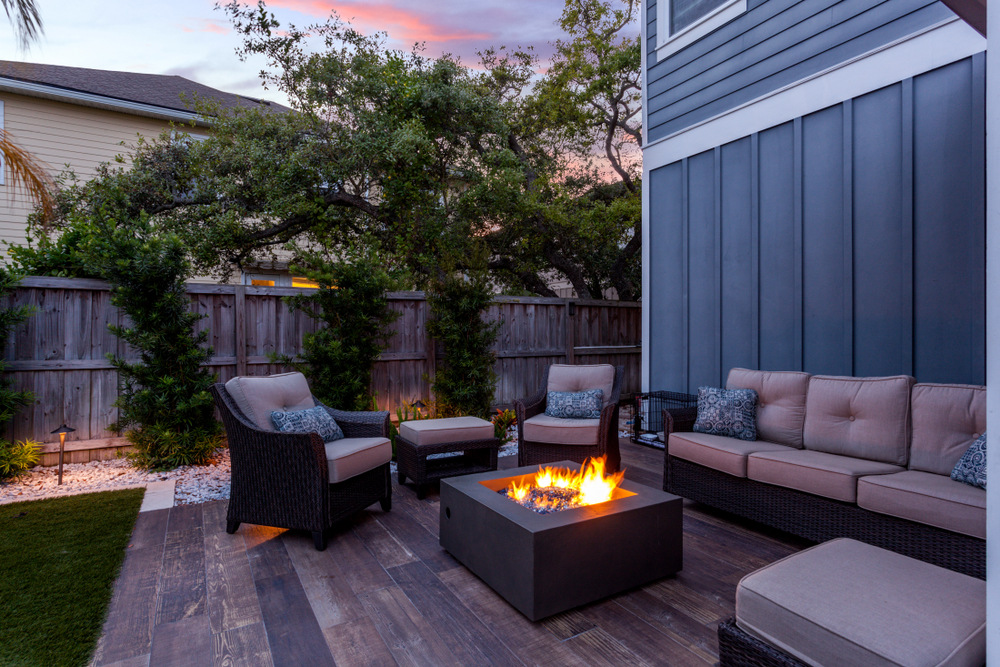 A cozy backyard patio with wicker chairs and sofas around a modern fire pit, set on wooden decking and surrounded by plants and a wooden fence at sunset.