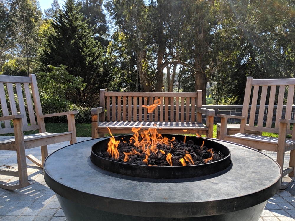 A fire pit with bright orange flames burns in a patio area, surrounded by wooden bench seating and rocking chairs, with trees and greenery in the background.