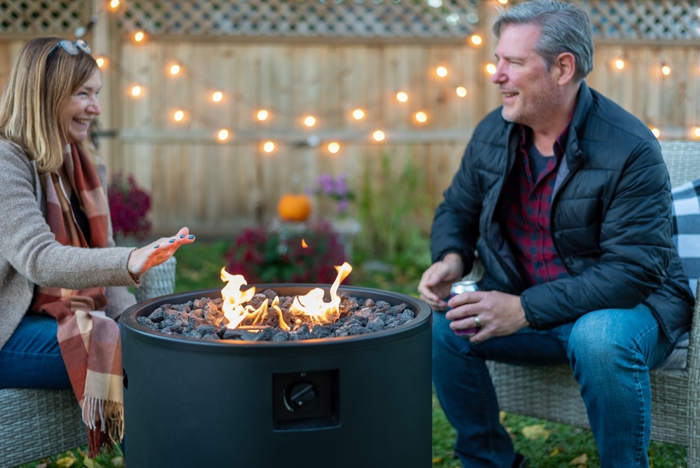 A woman and a man sit outdoors by a round fire pit, smiling and talking. String lights hang on a wooden fence in the background, creating a cozy, warm atmosphere in the backyard.