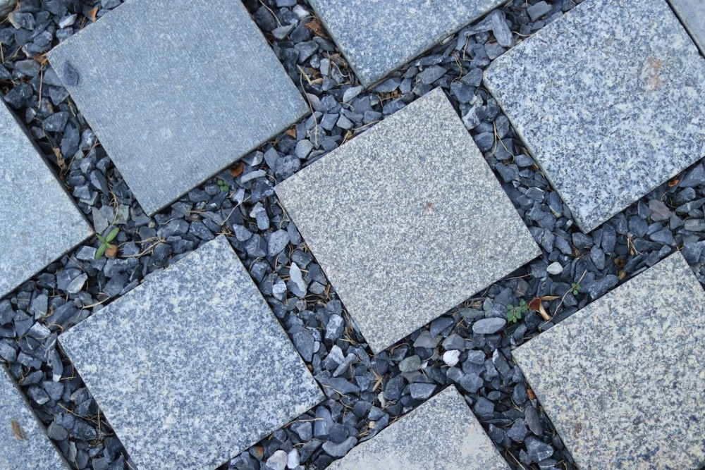 Gray square paving stones are arranged in a grid pattern, separated by small dark gravel stones filling the gaps between them. Some green plants are visible in the gravel.