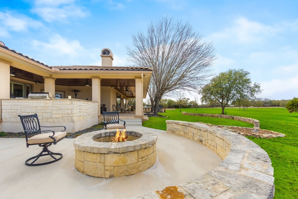 A patio with stone seating and a fire pit in the foreground, two chairs, and a covered outdoor area attached to a house, set in a large grassy yard with trees under a blue sky.