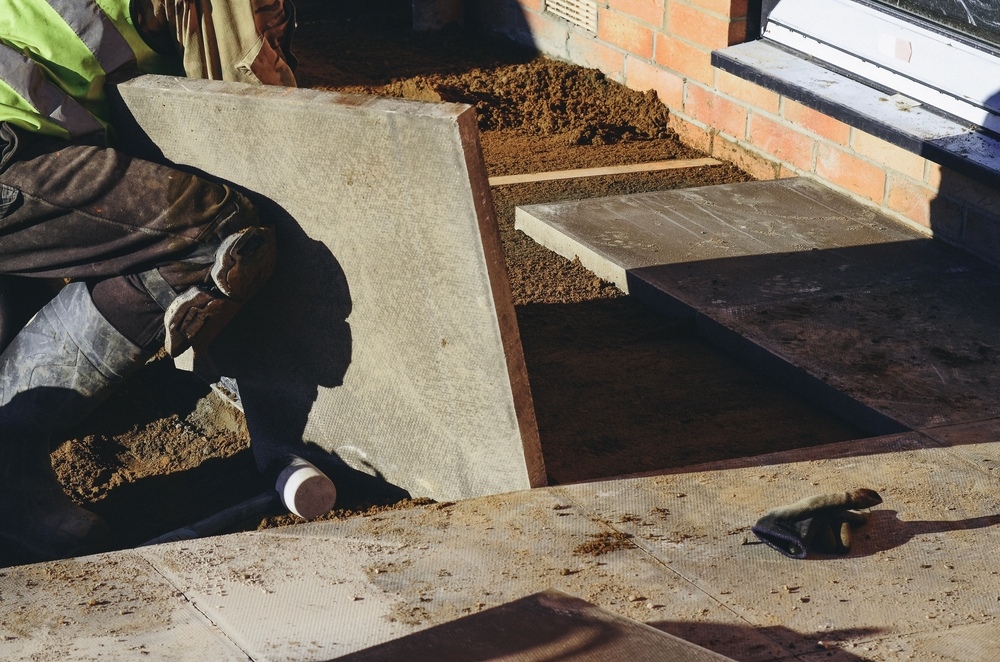 A construction worker in boots and workwear positions a large paving slab onto a sand base outside a brick building, with other slabs and a glove visible nearby.