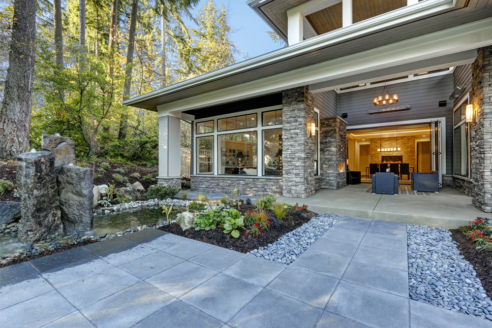 Modern house exterior with large windows, stone columns, open patio, and landscaped garden featuring rocks, a water feature, and lush greenery surrounded by tall trees.