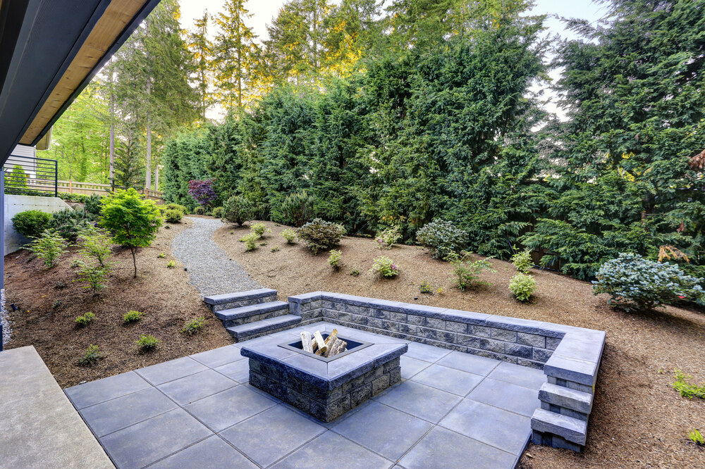 Modern backyard patio with a built-in stone fire pit, surrounded by low retaining walls and steps, bordered by a gravel path, young shrubs, and tall trees providing lush, green privacy.