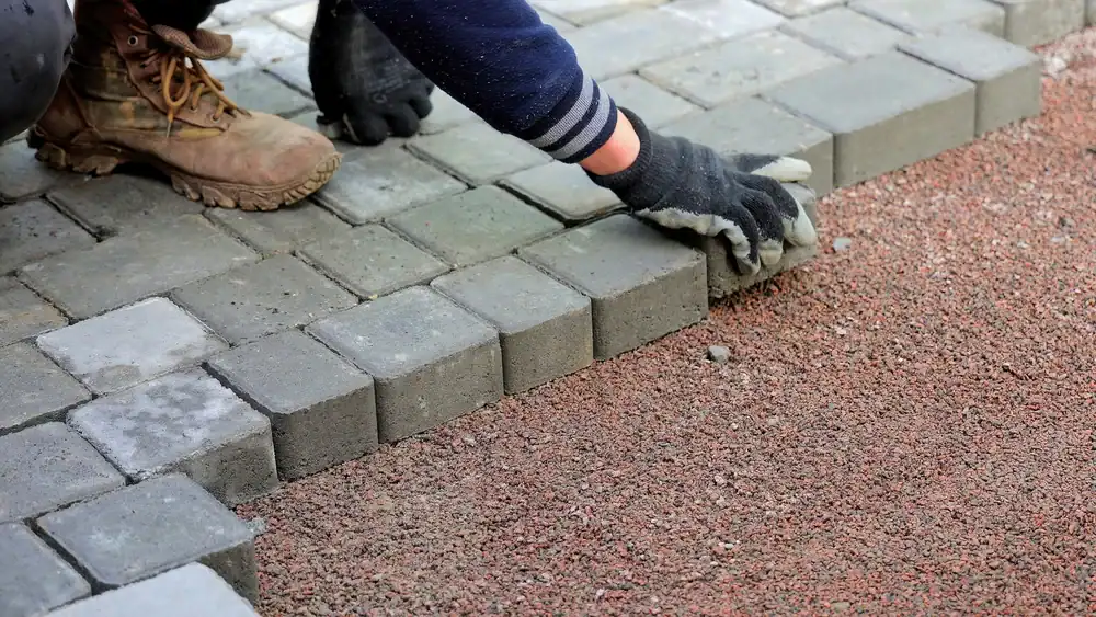 A masonry contractor in Suffolk County, NY, wearing work gloves and boots, is laying rectangular paving stones on a gravel surface, carefully placing each stone to form a precise pattern.