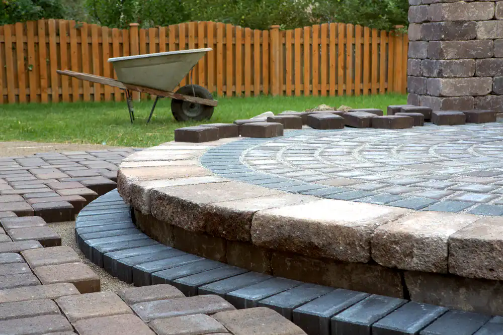 A partially completed circular stone patio with various paving stones laid out; a wheelbarrow sits on grass near a wooden fence in the background, showcasing the quality work of a masonry contractor Suffolk County, NY.