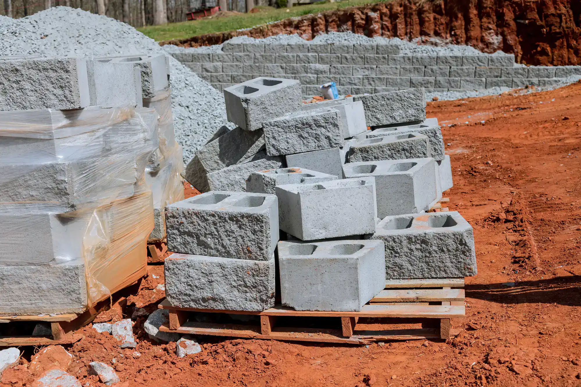 A stack of concrete cinder blocks is arranged on wooden pallets at a construction site with red soil and gravel. Wrapped blocks and a partially built stone retaining wall are visible in the background.