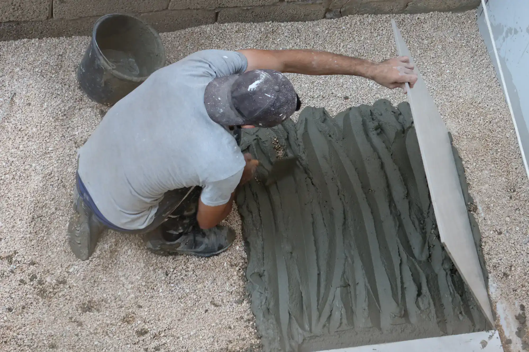 A construction worker seen from above spreads and levels wet cement with a tool on a gravel surface, wearing a grey shirt, dark pants, and a cap. A bucket sits nearby.