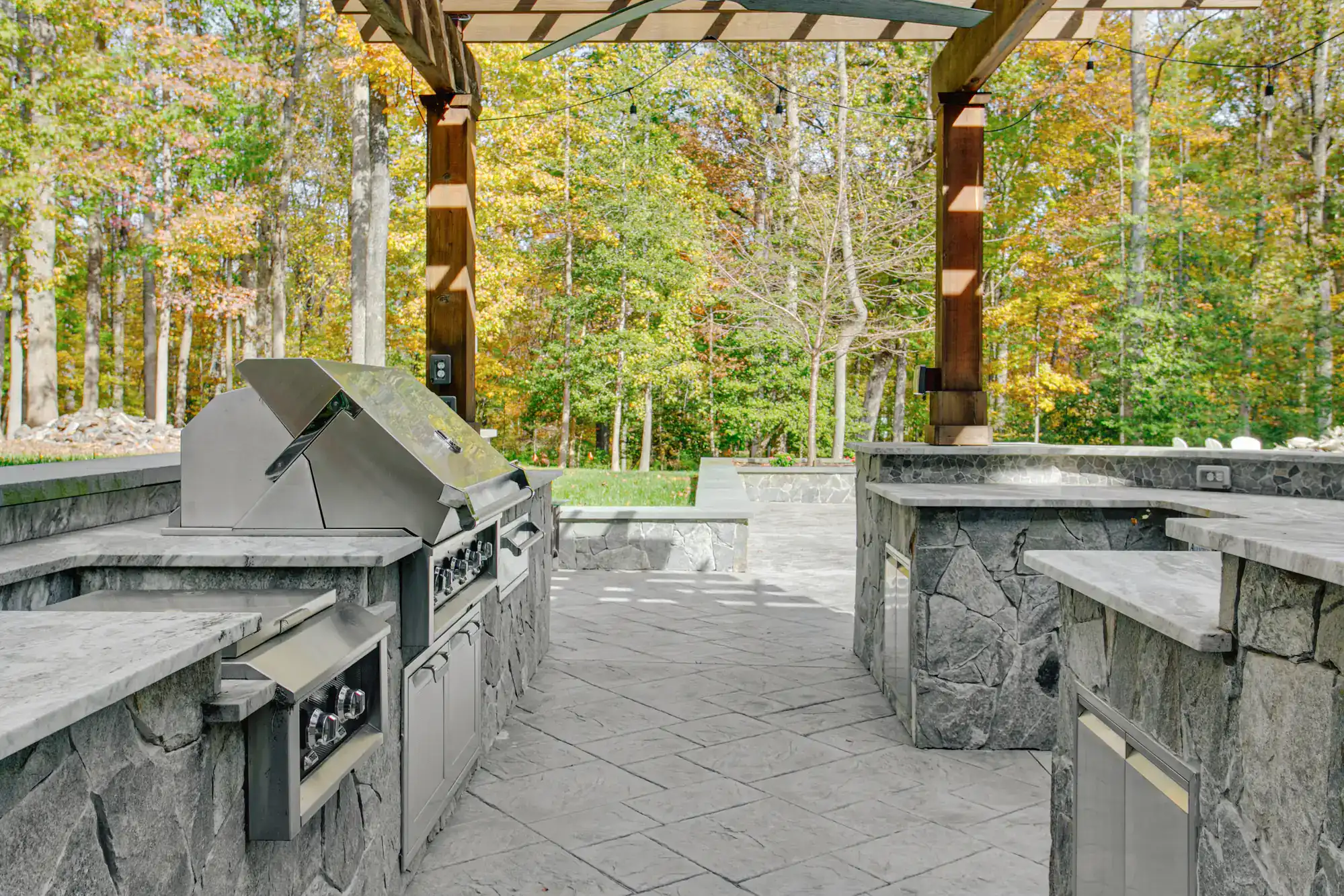 Outdoor kitchen with stainless steel grill and appliances, stone countertops, and a pergola. Surrounded by trees with autumn foliage, the kitchen sits on a stone patio with ample counter space and natural light.
