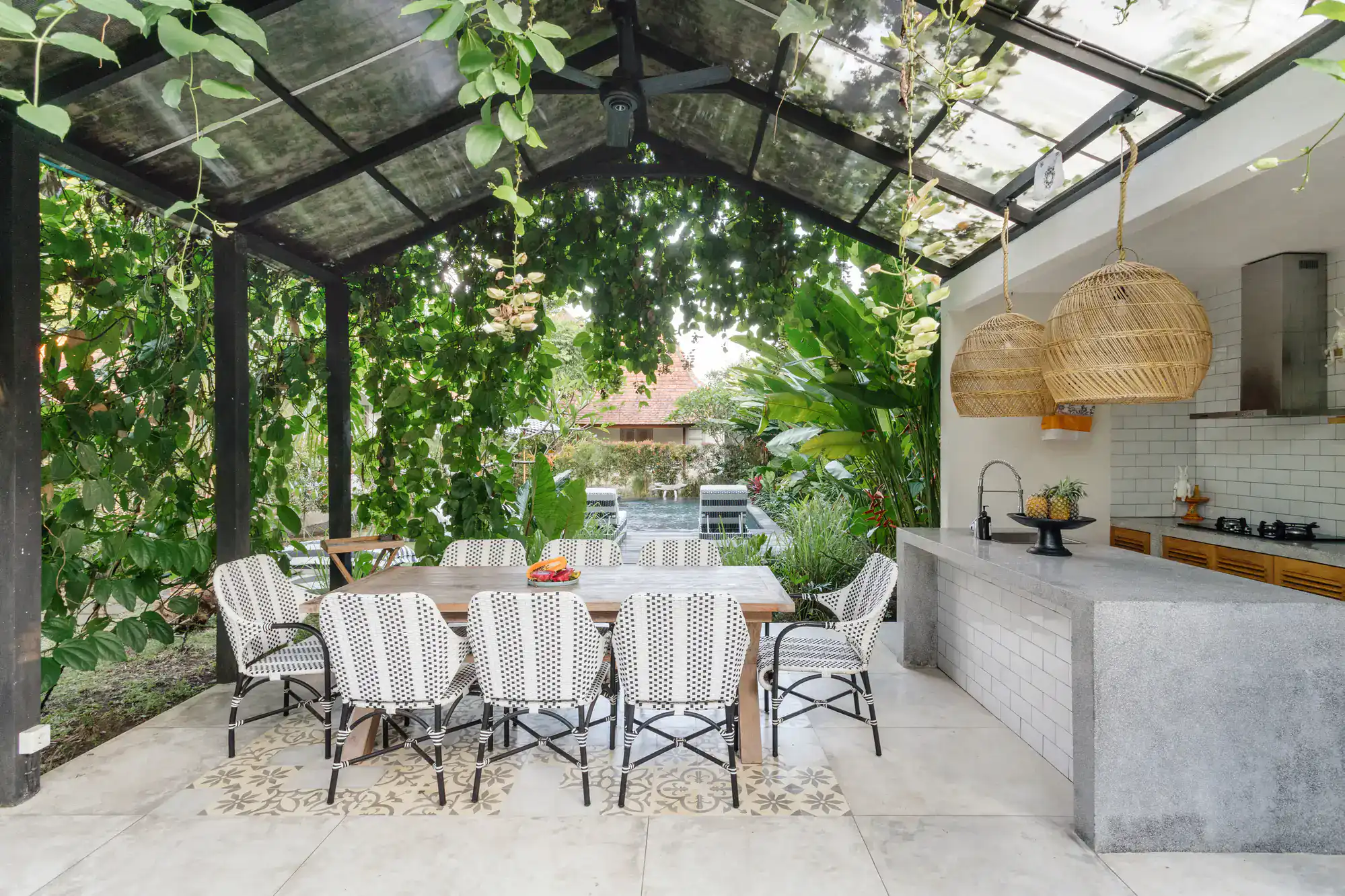 A stylish outdoor dining area with a wooden table, patterned chairs, hanging wicker lamps, and a modern kitchen under a glass-roofed patio, surrounded by lush green plants and trees.