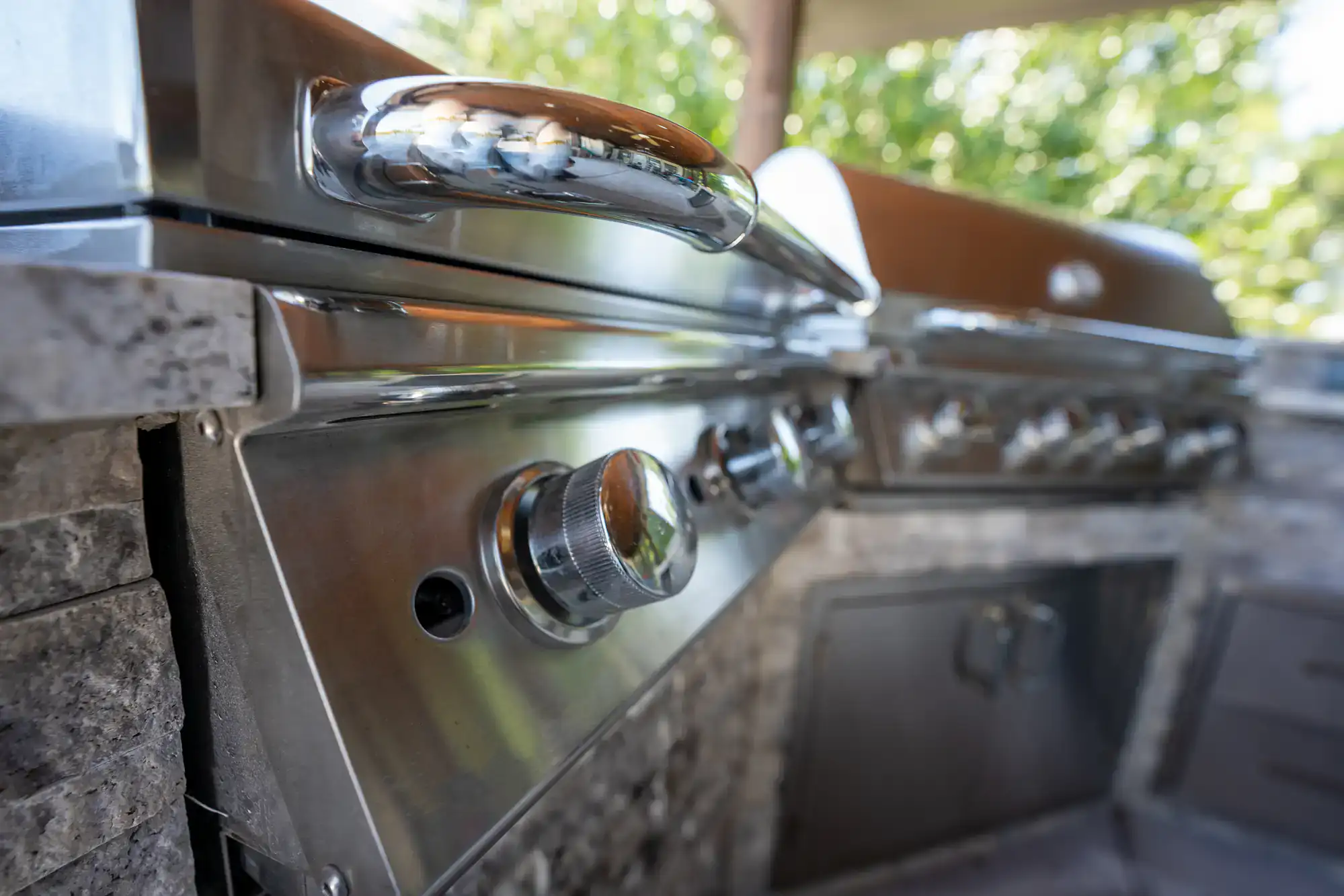 Close-up of shiny stainless steel knobs and handles on an outdoor gas grill, with stone countertops and blurred greenery in the background.