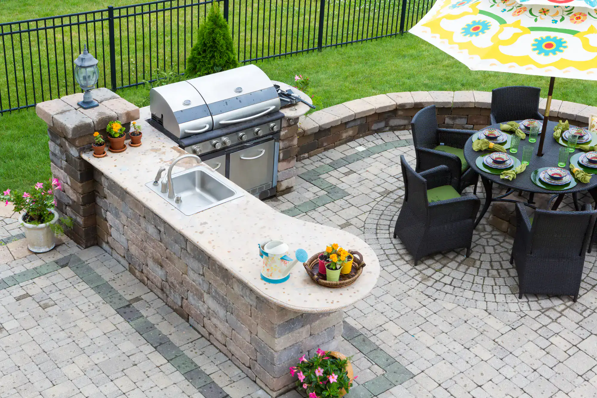 Outdoor patio with a stone kitchen island featuring a grill, sink, potted plants, and decorative items. Nearby is a round table set for six under a large patterned umbrella, with green grass and a black fence in the background.