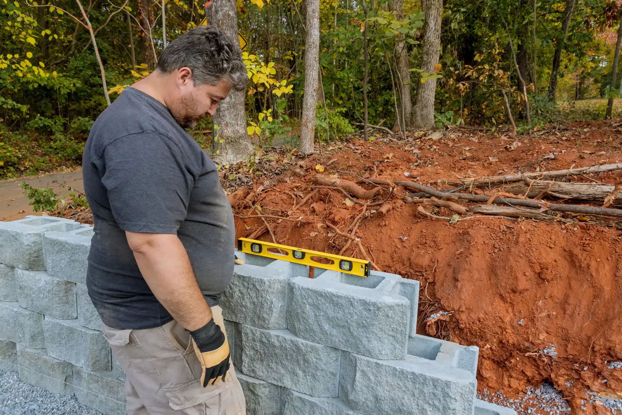 A man wearing gloves checks the level of a gray stone retaining wall with a yellow spirit level, next to a dirt embankment and forested area.