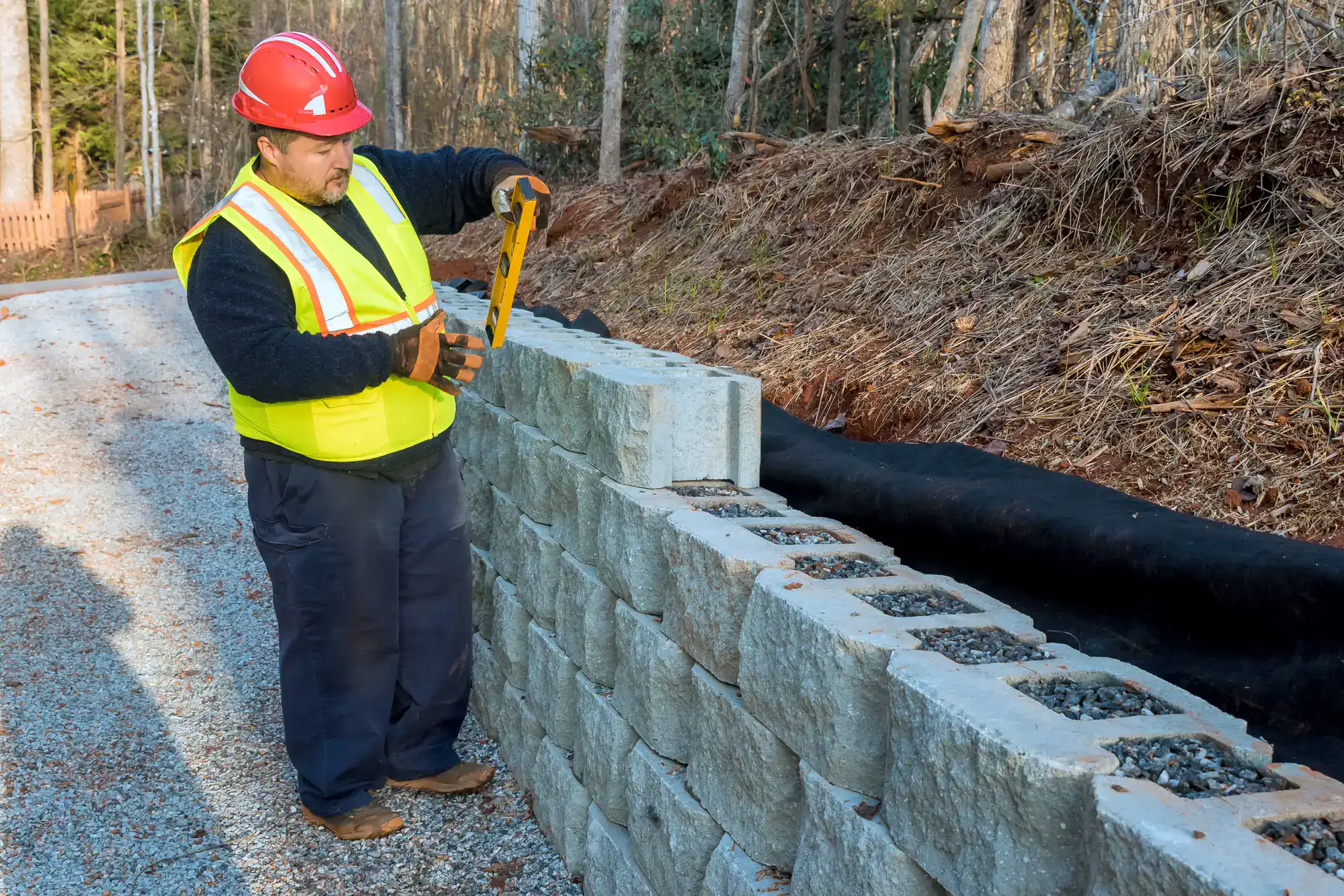 A construction worker in a yellow safety vest and red helmet uses a level to check concrete blocks while building a retaining wall next to a gravel road and a slope with trees.