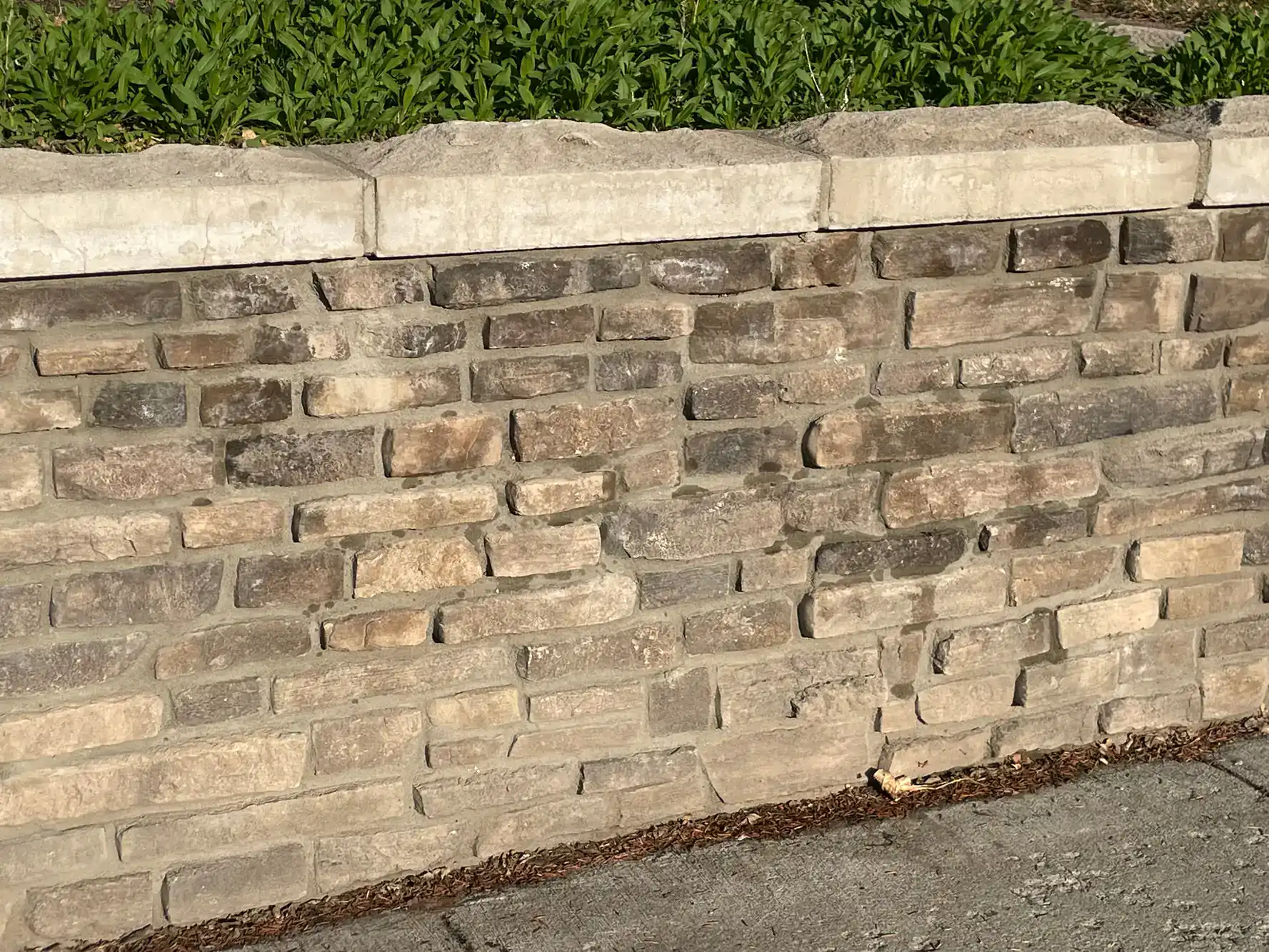 A stone retaining wall made of rectangular, tan and brown bricks supports a raised bed with green plants. Sunlight casts shadows on the textured wall and gray sidewalk below.